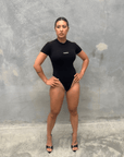 Woman wearing a black T-shirt short sleeve bodysuit standing against a grey concrete wall front of bodysuit
