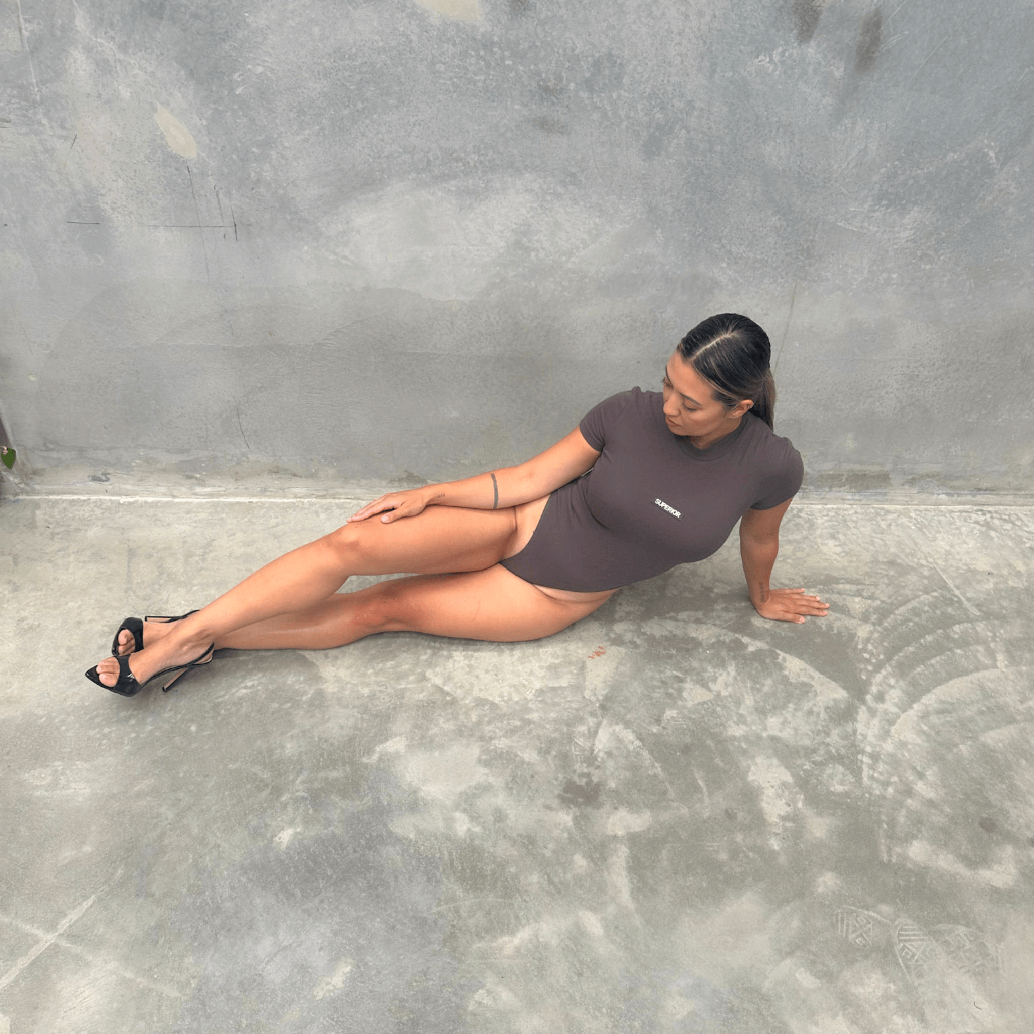 Woman in a charcoal short sleeve bodysuit laying on the floor against a grey wall front on
