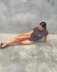 Woman in a charcoal short sleeve bodysuit laying on the floor against a grey wall front on