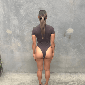 Woman in a charcoal short sleeve bodysuit standing against a grey wall showing back of the bodysuit