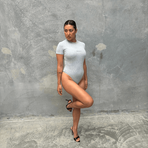 Grey short sleeve bodysuit modelling front of grey concrete wall side on