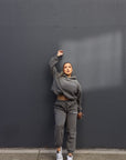 posing against charcoal wall, one arm up in the air in the full dark grey set