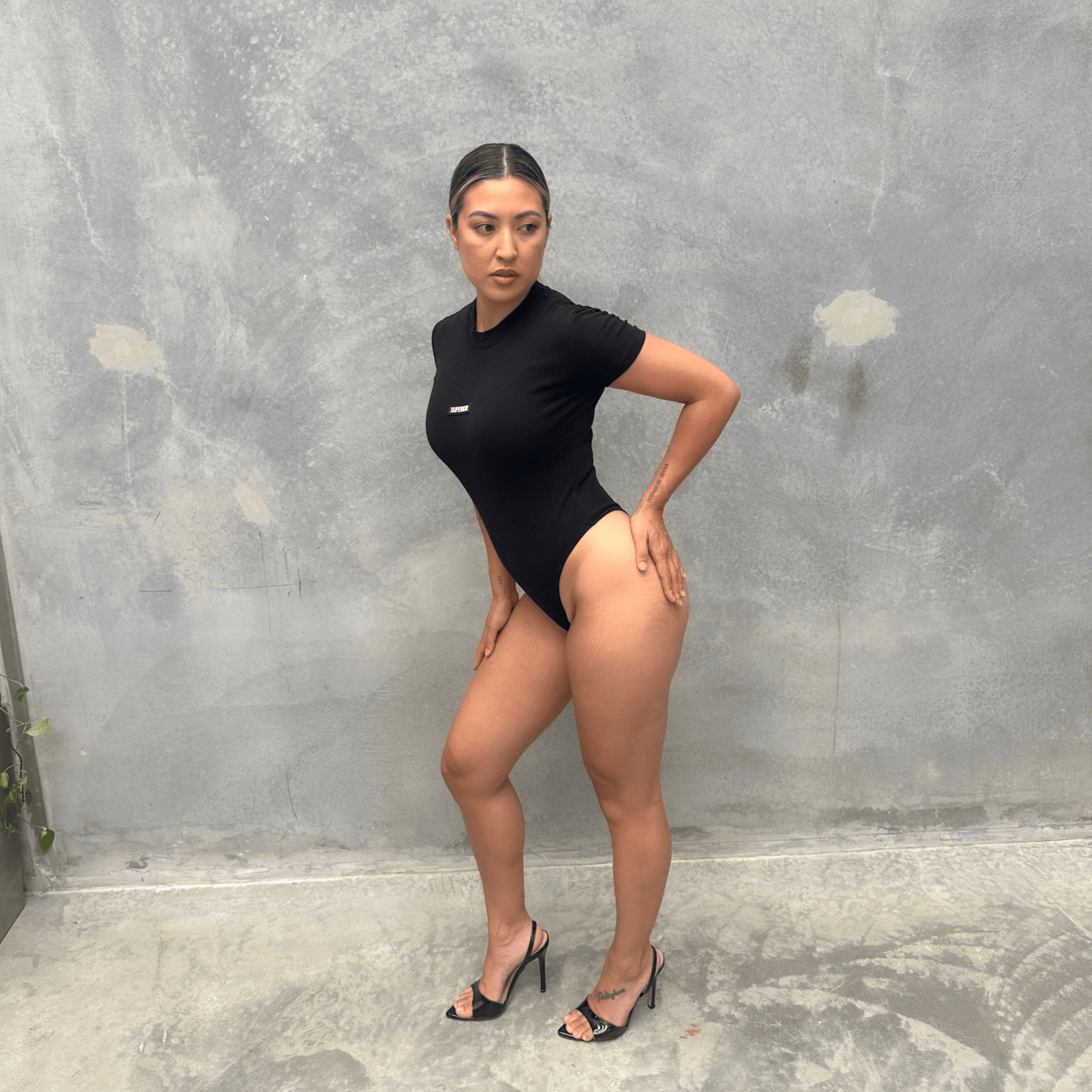 Woman in a black short sleeve bodysuit against a grey concrete wall posing from the side 