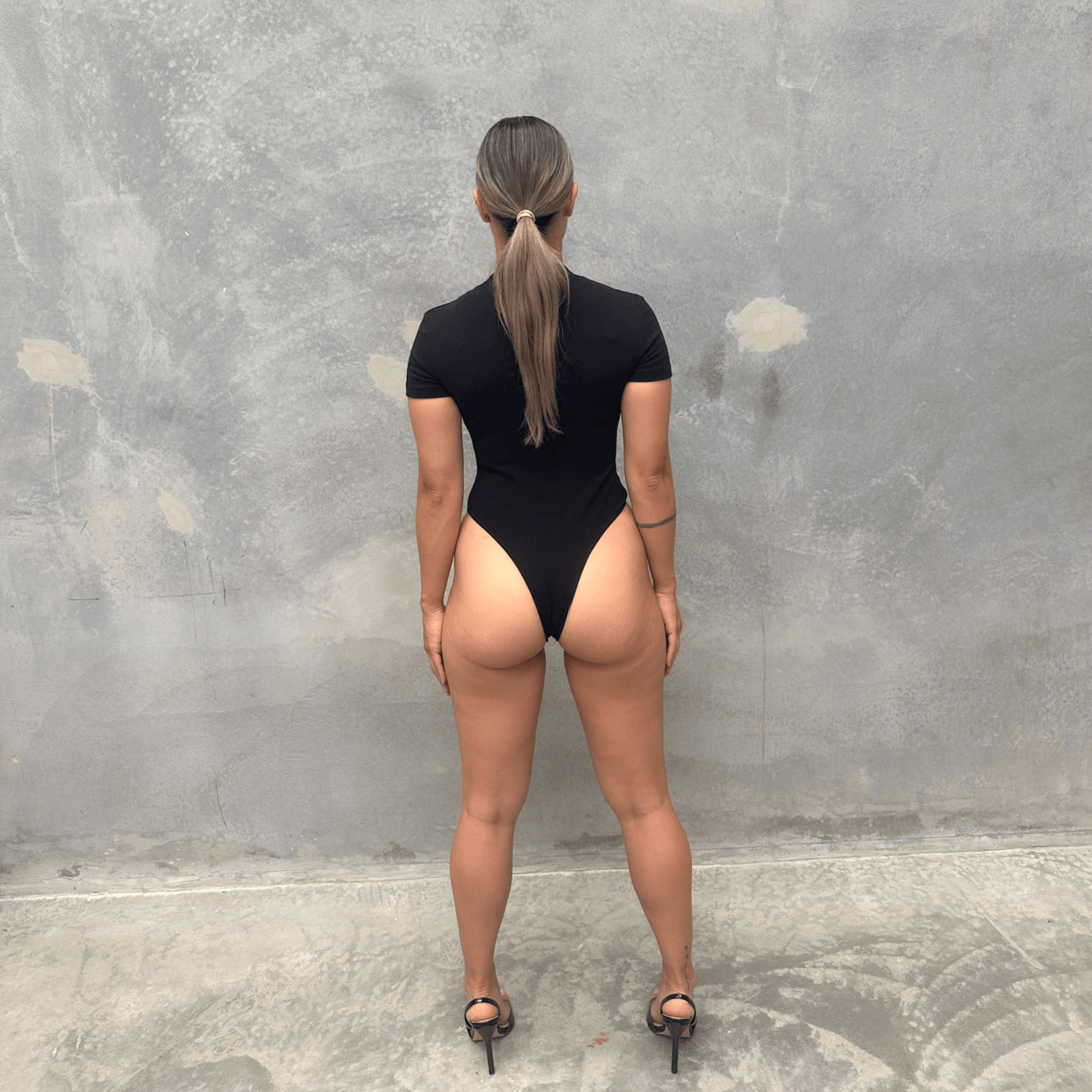Woman in a black short sleeve bodysuit against a grey concrete wall posing from the back 