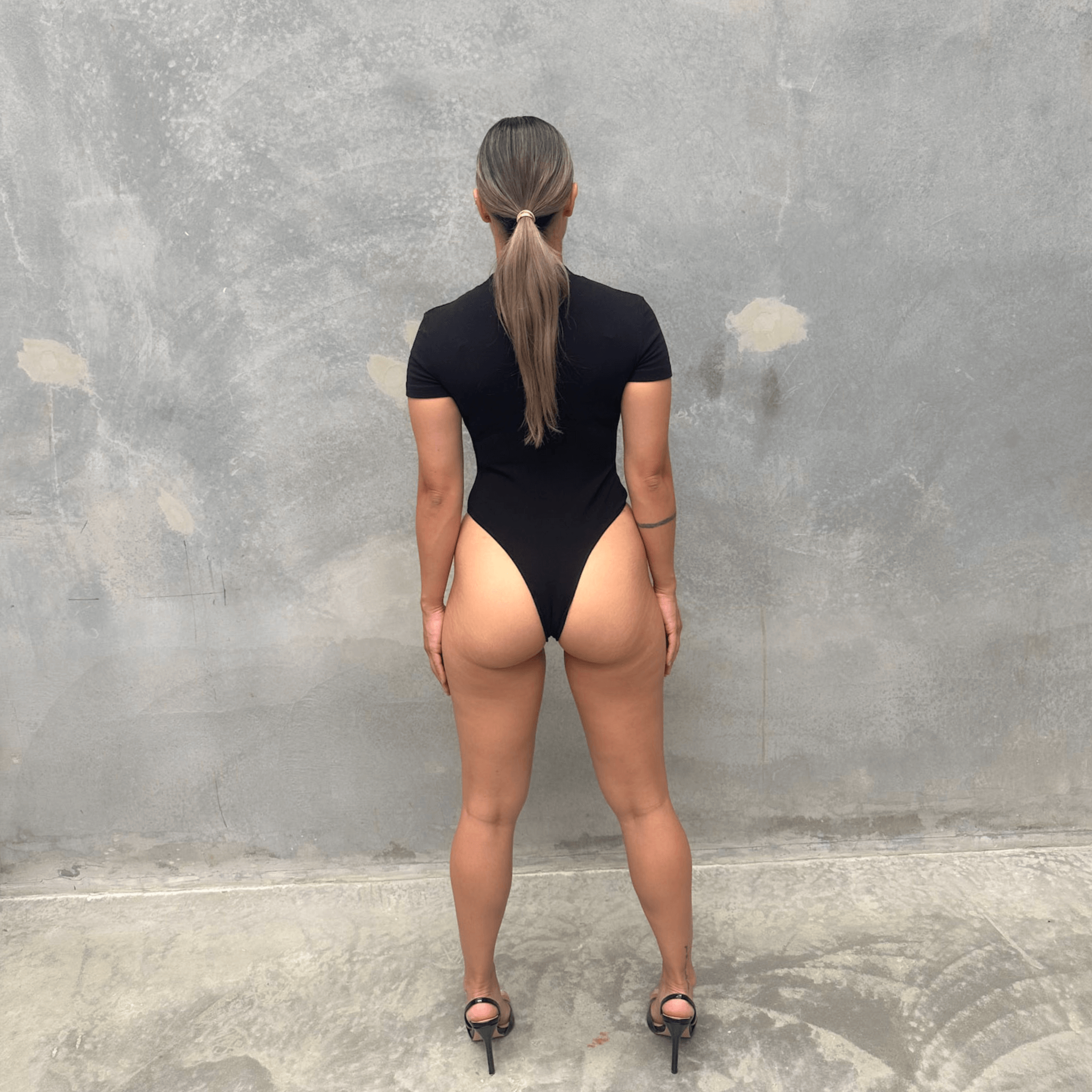 Woman in a black short sleeve bodysuit against a grey concrete wall posing from the back 