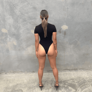 Woman in a black short sleeve bodysuit against a grey concrete wall posing from the back 