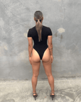 Woman in a black short sleeve bodysuit against a grey concrete wall posing from the back 