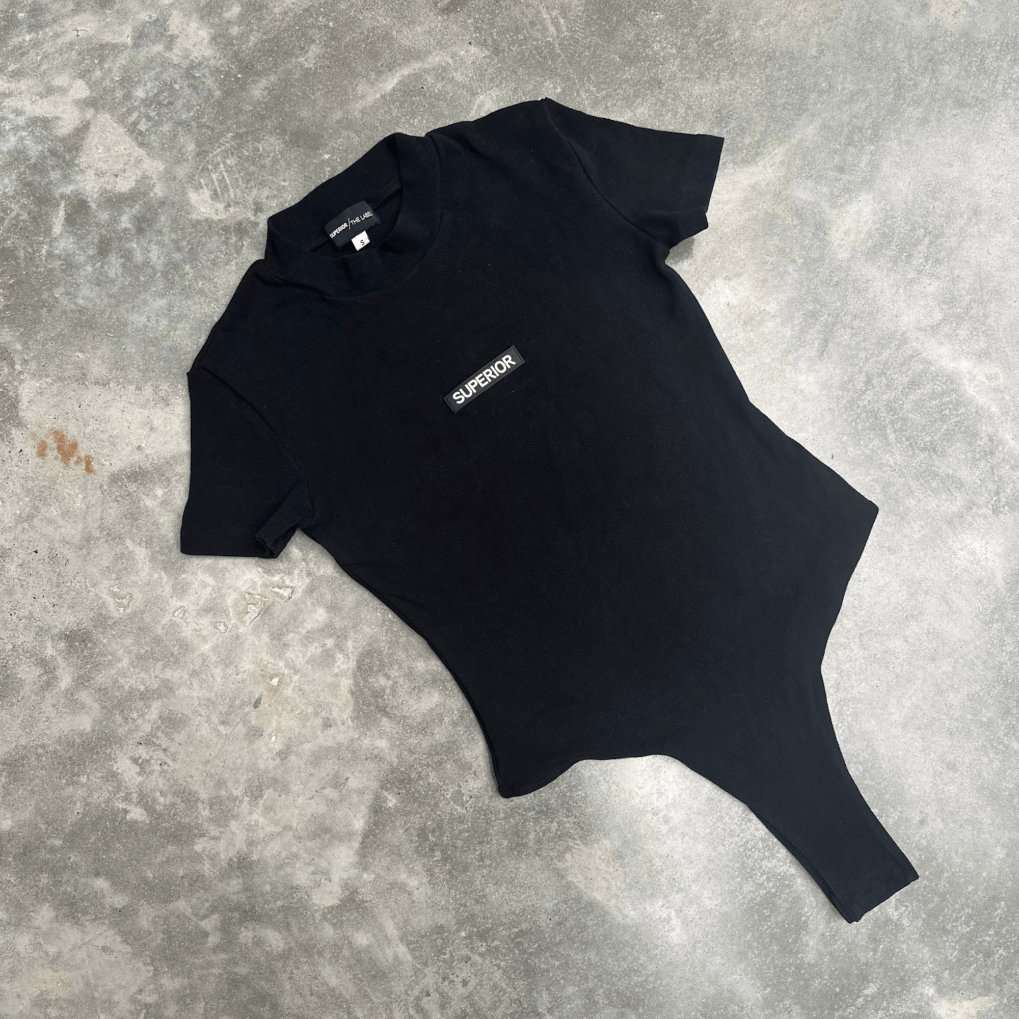 Black t-shirt short sleeve bodysuit with ‘SUPERIOR’ logo on a grey concrete surface for flatlay product shot 