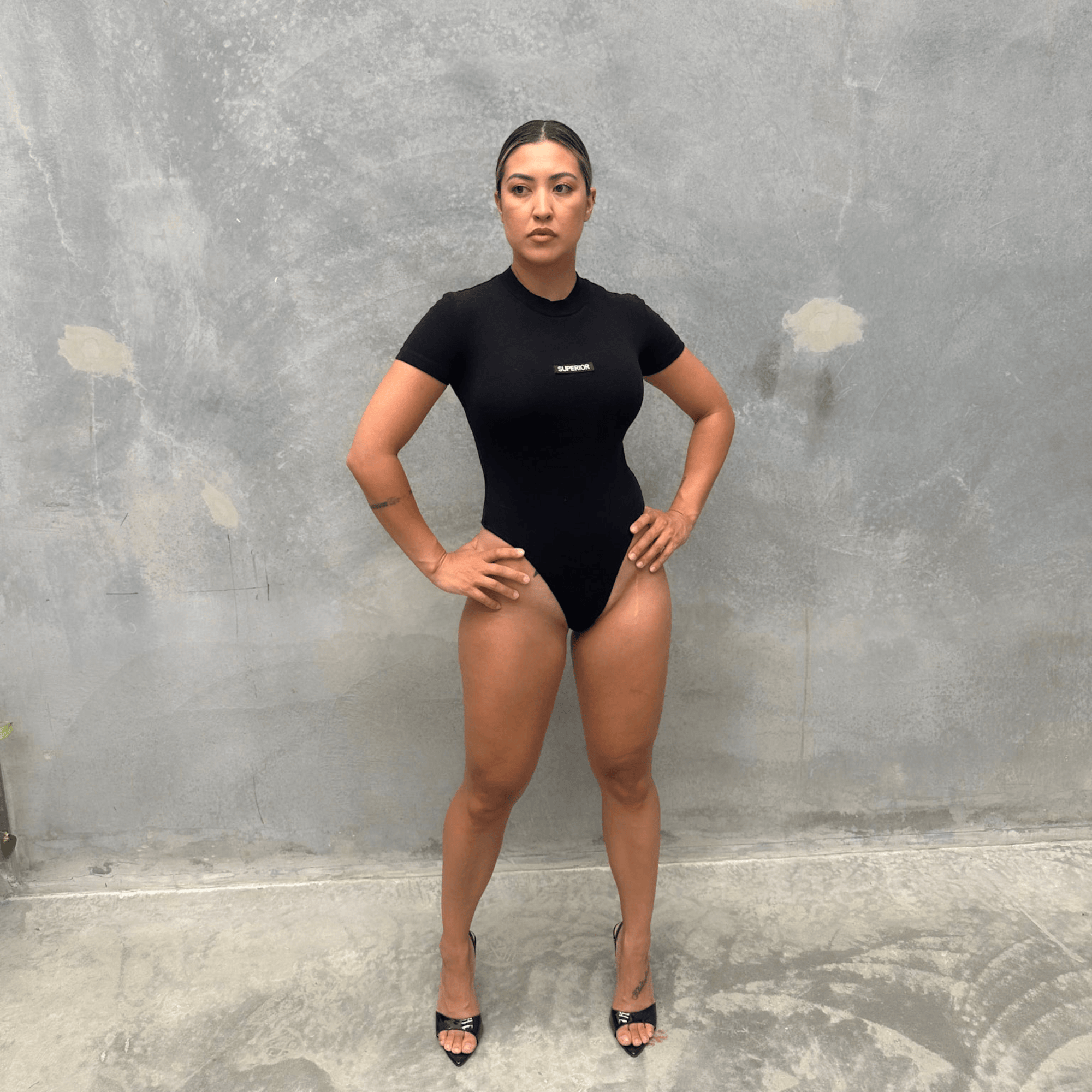 Woman wearing a black T-shirt short sleeve bodysuit standing against a grey concrete wall front of bodysuit 