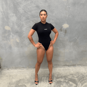 Woman wearing a black T-shirt short sleeve bodysuit standing against a grey concrete wall front of bodysuit 