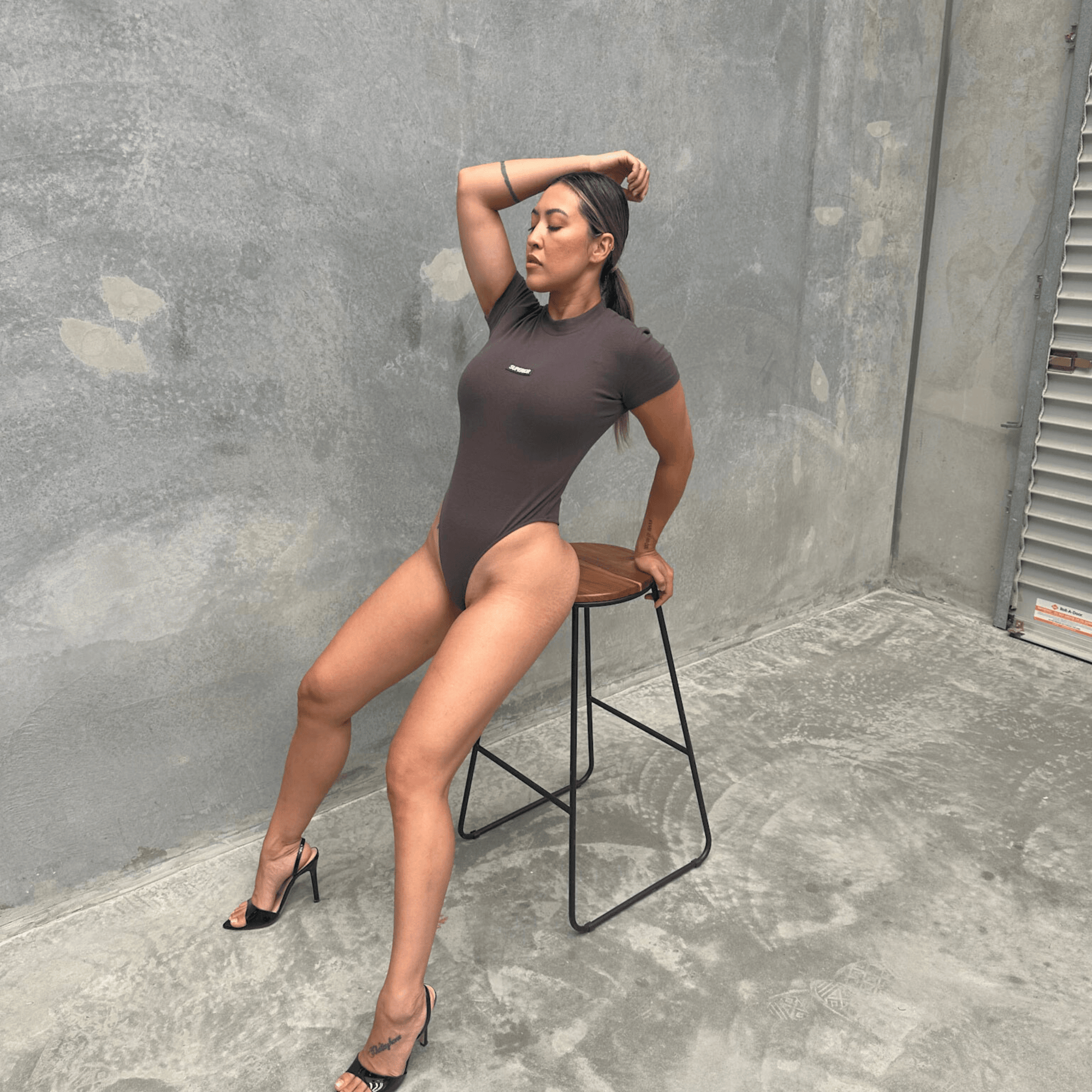 Woman in a charcoal short sleeve bodysuit sitting on a stool against a grey wall