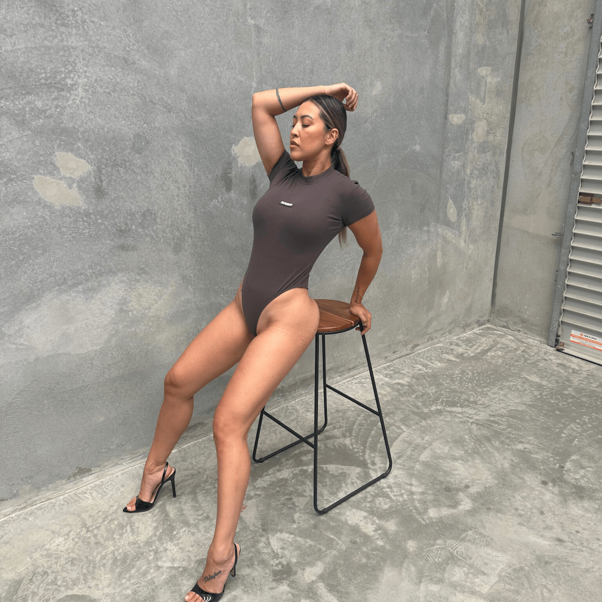 Woman in a charcoal short sleeve bodysuit sitting on a stool against a grey wall