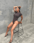 Woman in a charcoal short sleeve bodysuit sitting on a stool against a grey wall