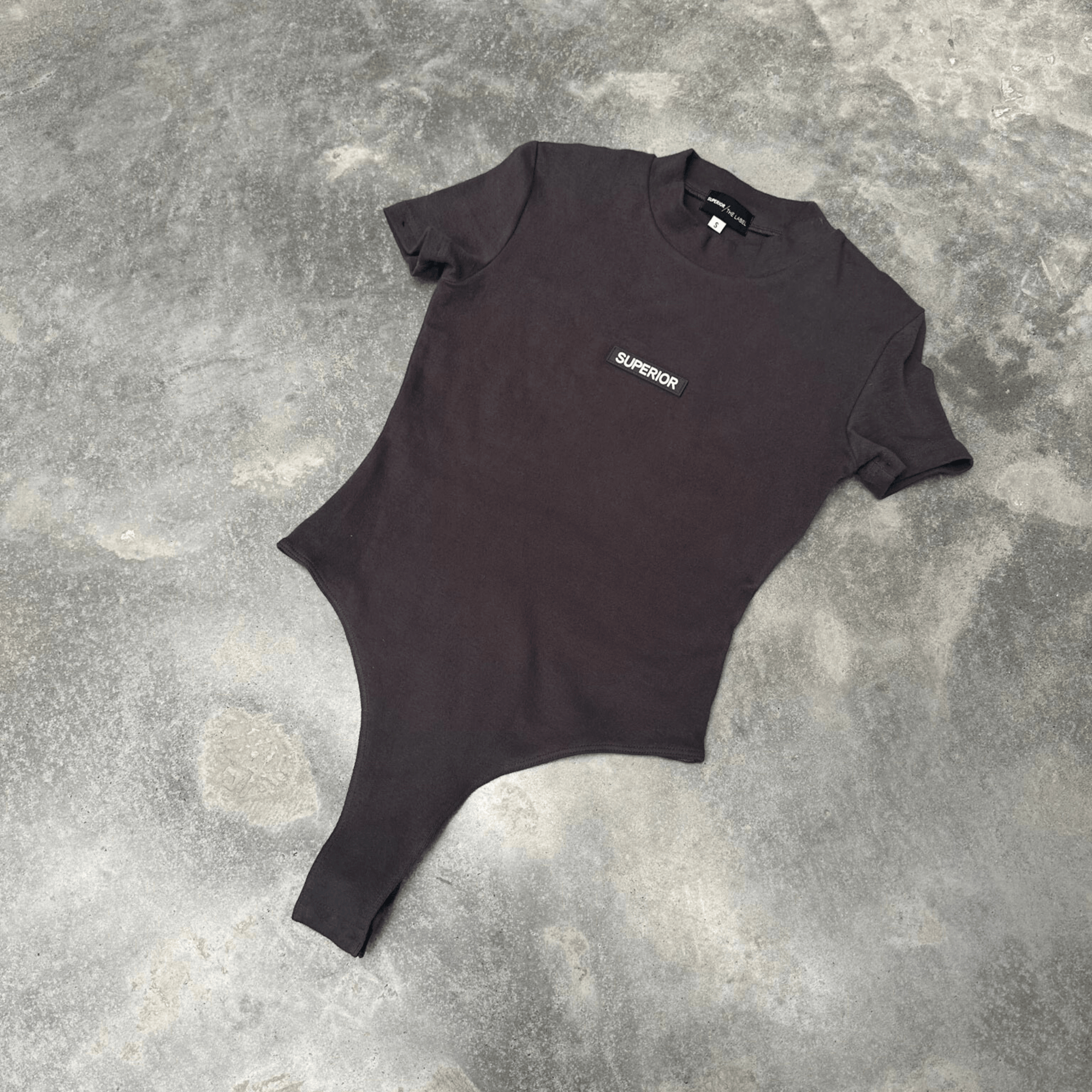 Black t-shirt short sleeve bodysuit with 'SUPERIOR' logo on a textured gray surface front on in flat lay position 